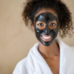 Smiling young woman with charcoal face mud looking at camera isolated on beige background with copy space. Portrait of african woman with curly hair wearing white bath robe getting black purifying mask at spa. Happy girl feeling relaxed at salon after face beauty facial treatment with peeling scrub clay.